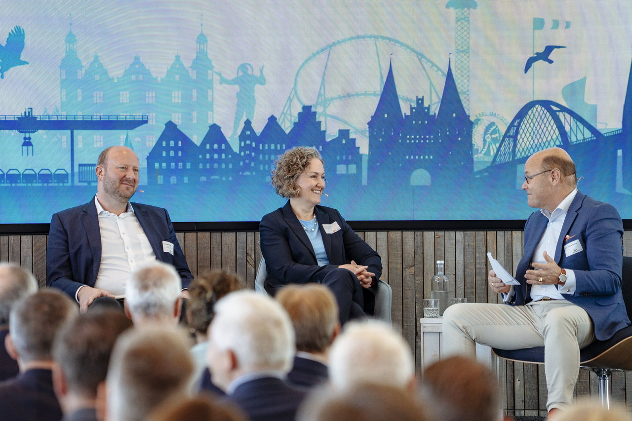 Drei Diskutierende auf Sesseln vor einer Leinwand mit Lübecker Stadtsilhouette
