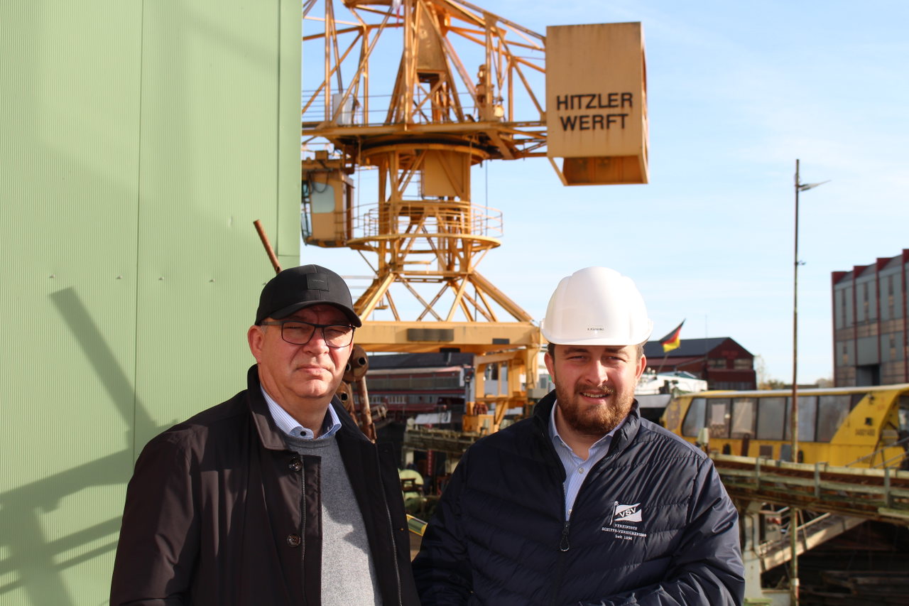 Marek Klimenko mit Mütze und Kai Klimenko mit weißem Schutzhelm vor einem gelben Werftkran mit der Aufschrift Hitzler Werft