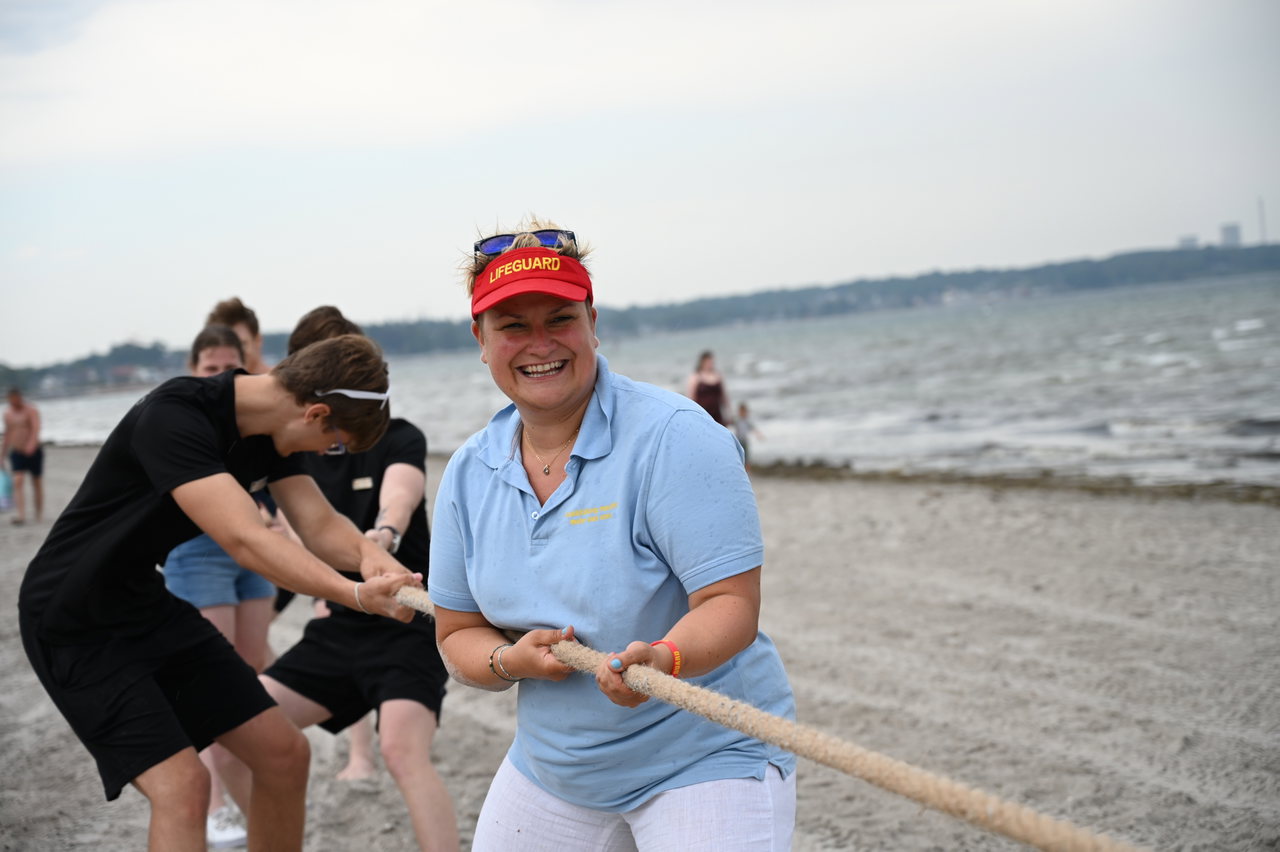 Eine Polizistin mit roter Kappe und Jugendliche beim Tauziehen am Strand