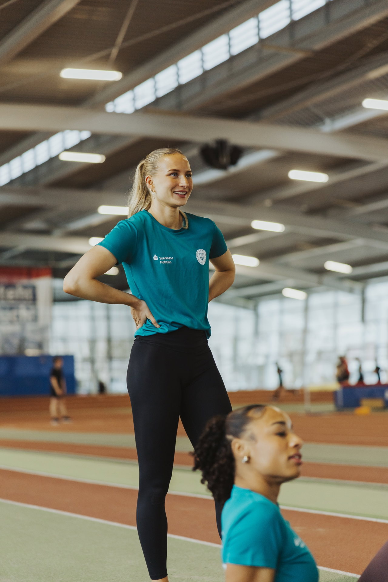 Leichtathletin im türkisfarbenen Shirt steht lächelnd in der Halle, im Vordergrund eine Teamkollegin