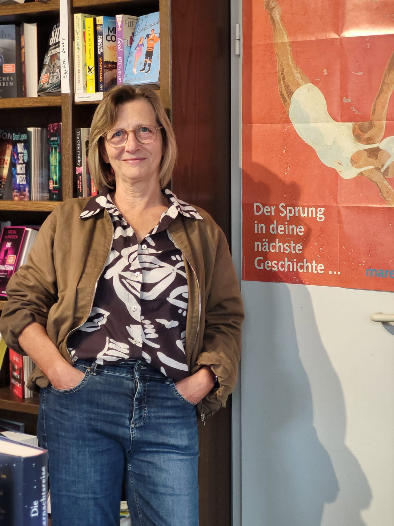 Ulrike Herberg mit Brille und brauner Jacke vor einem Bücherregal in ihrer Buchhandlung