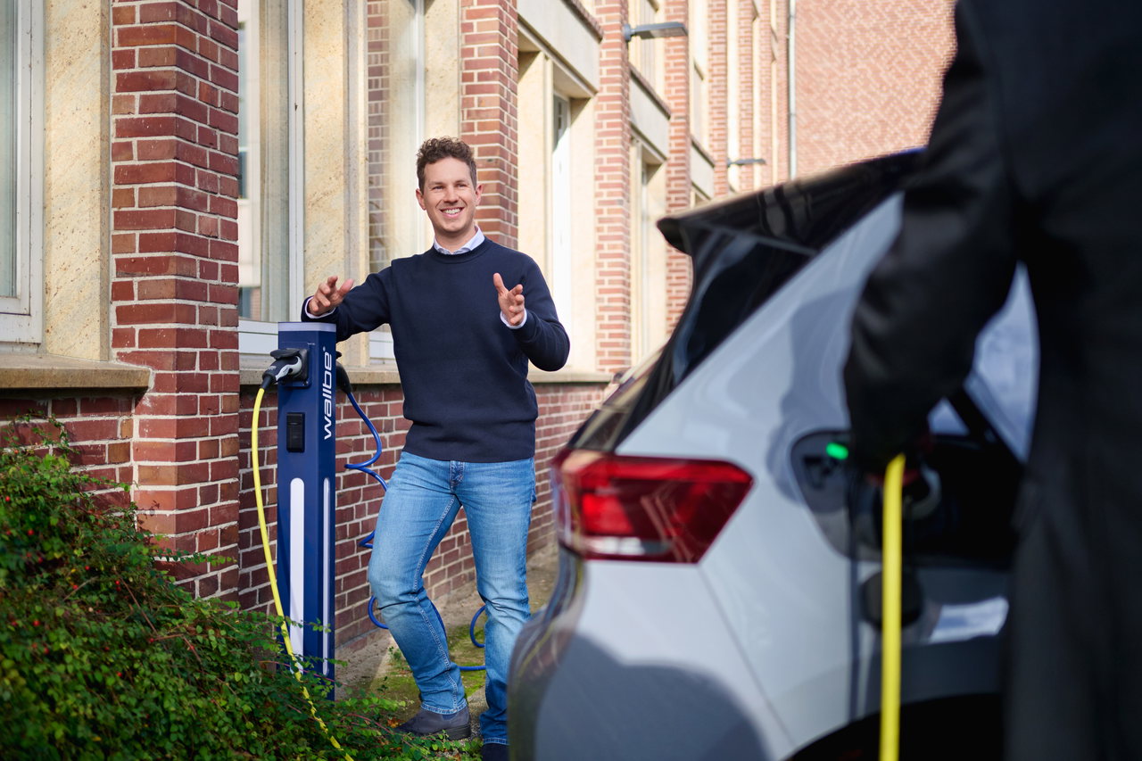 Jurek Schwekendiek im dunklen Pullover an einer Ladesäule vor einem Backsteingebäude, im Vordergrund wird ein Auto geladen