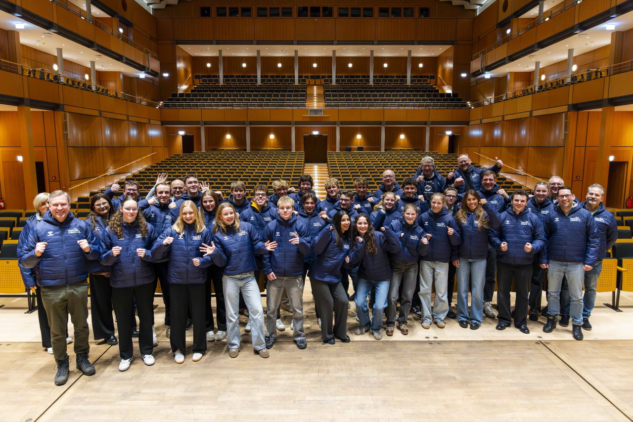 Gruppenfoto des Team Lübeck in blauen Jacken auf einer Bühne vor leeren Zuschauerrängen