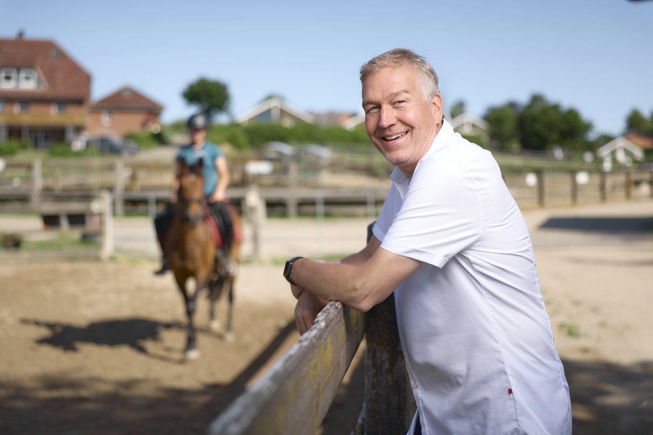 Lächelnder Mann in weißem Hemd lehnt am Zaun eines Reitplatzes, dahinter eine Reiterin auf einem Pferd