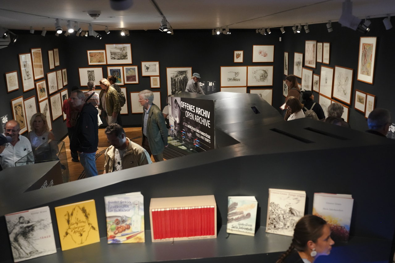 Blick von oben in die Ausstellung mit Zeichnungen an dunklen Wänden und Grass-Büchern im Vordergrund
