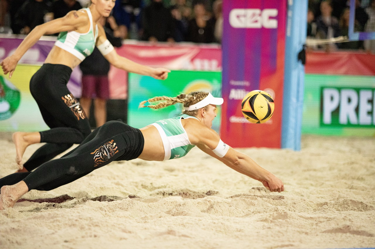 Beachvolleyballerin hechtet im Sand nach dem Ball, im Hintergrund ihre Teampartnerin und Zuschauer