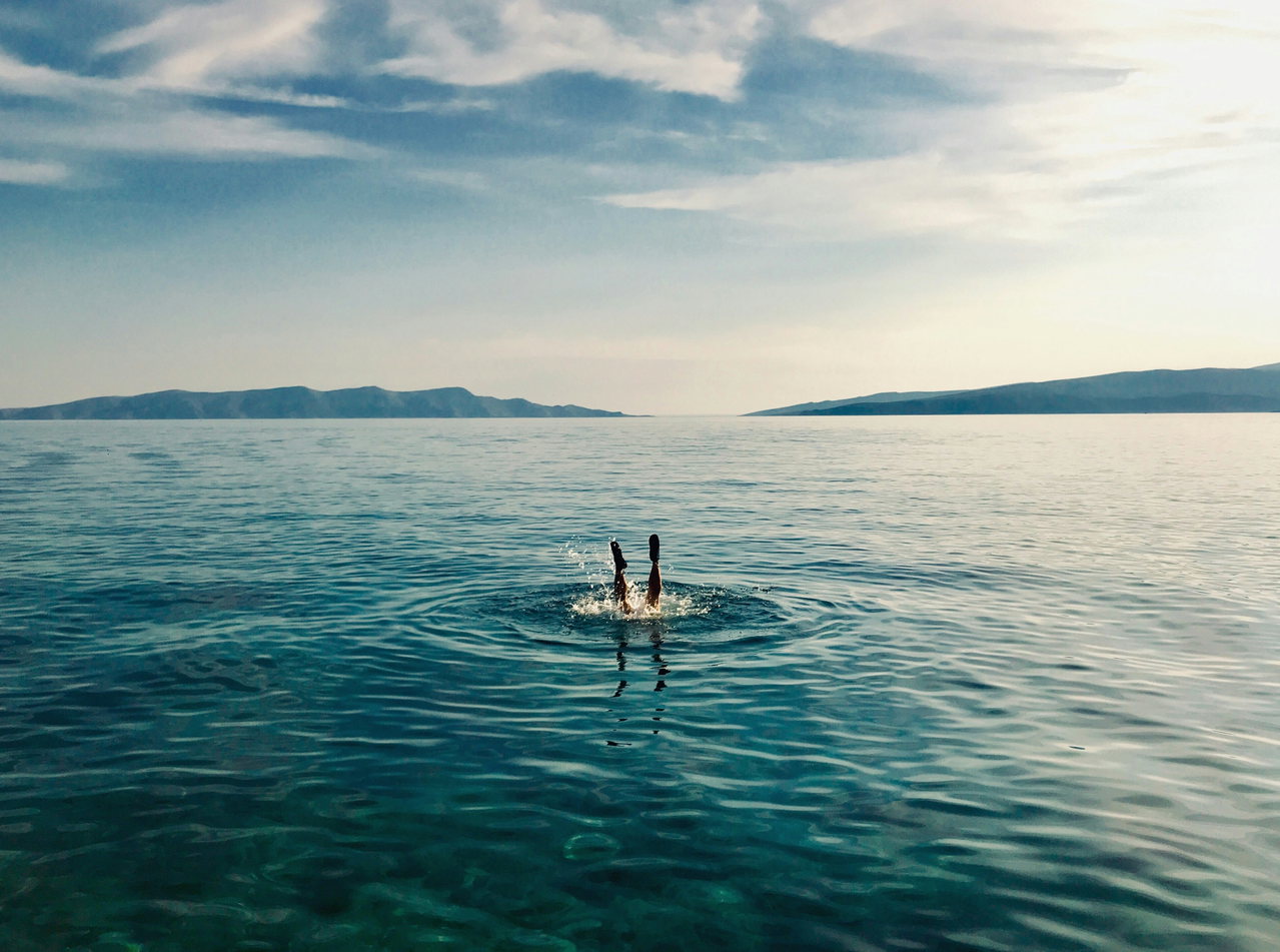 Zwei Beine ragen beim Abtauchen aus dem ruhigen Meer, im Hintergrund liegen Inseln