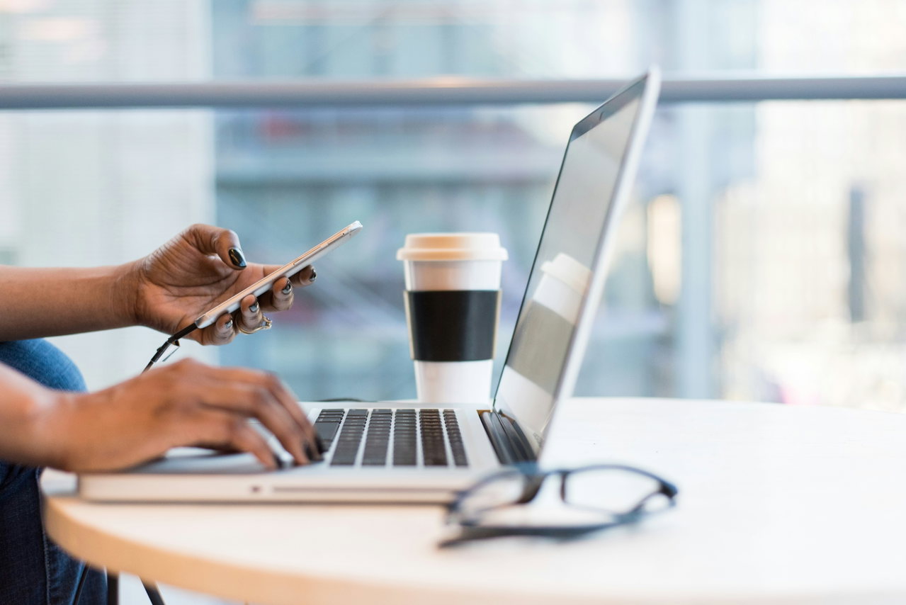 Hände an Laptop und Smartphone auf einem runden Tisch, daneben ein Kaffeebecher und eine Brille