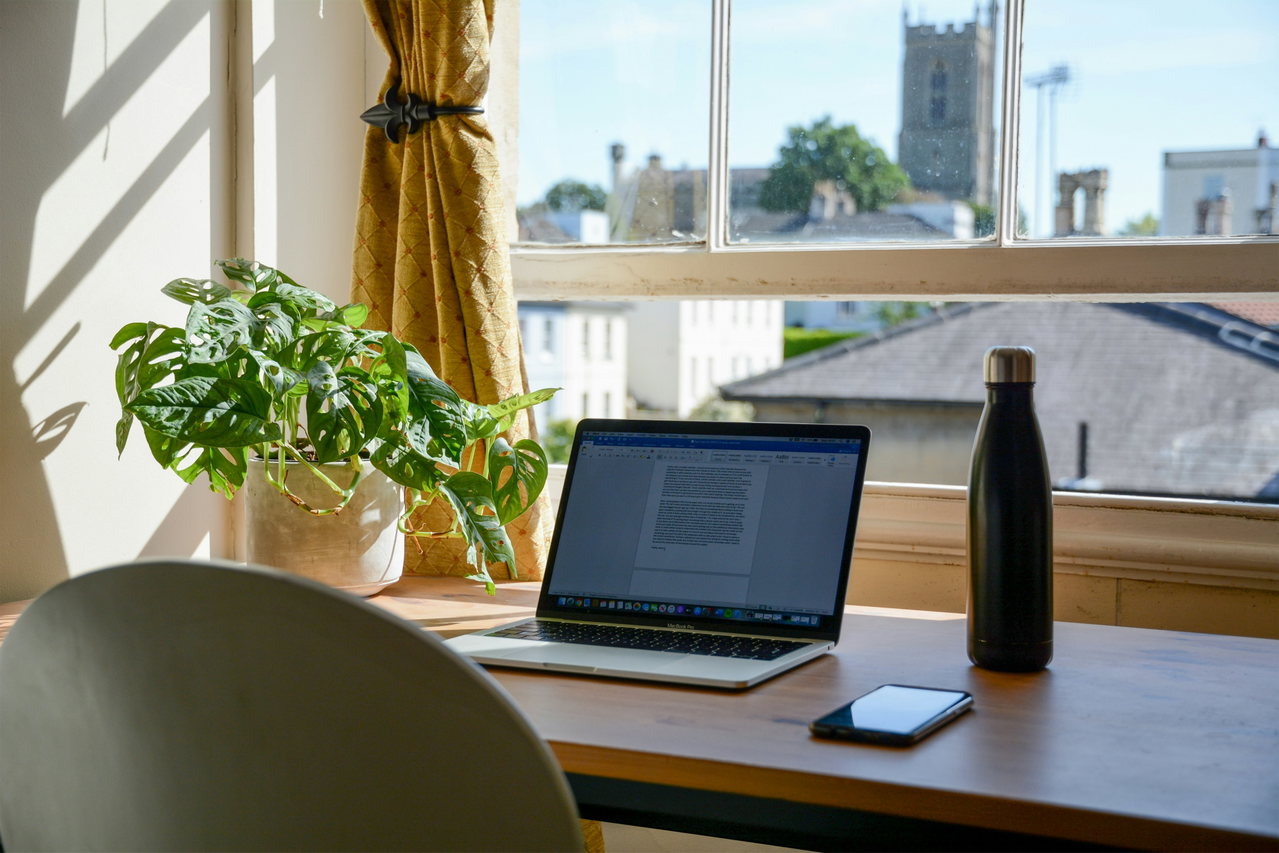 Aufgeräumter Arbeitsplatz am Fenster mit Laptop, Grünpflanze und Trinkflasche, draußen Dächer und ein Kirchturm