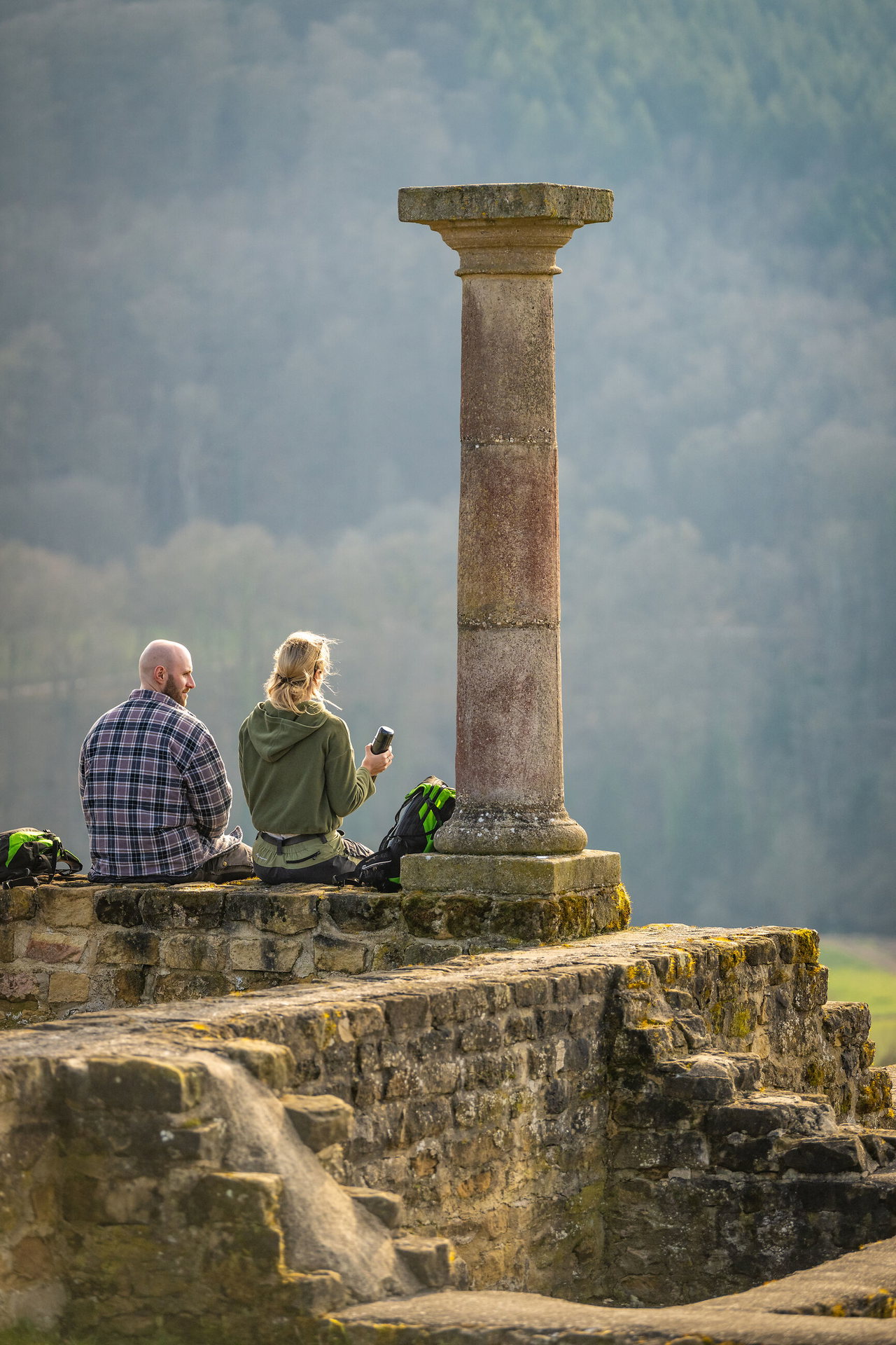 Zwei Personen sitzen auf einer steinernen Ruine neben einer einzelnen historischen Säule und blicken in die weite, sanft verschwommene Landschaft.