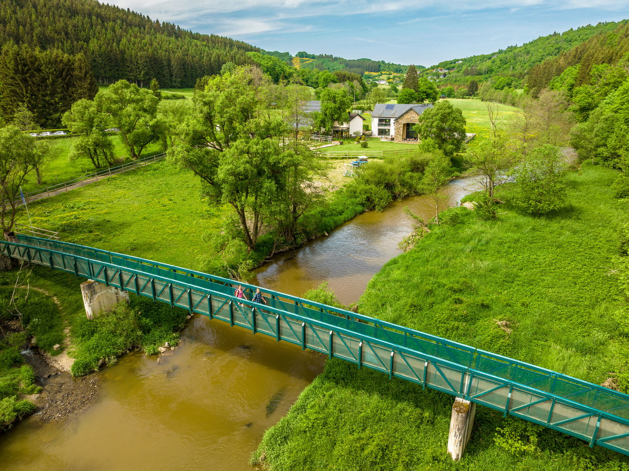 Eine grüne Tallandschaft mit einem Fluss, der unter einer langen türkisfarbenen Fußgängerbrücke hindurchfließt. Zwei Wandernde gehen über die Brücke, umgeben von Wiesen, Bäumen und sanften Hügeln mit einigen Häusern im Hintergrund.