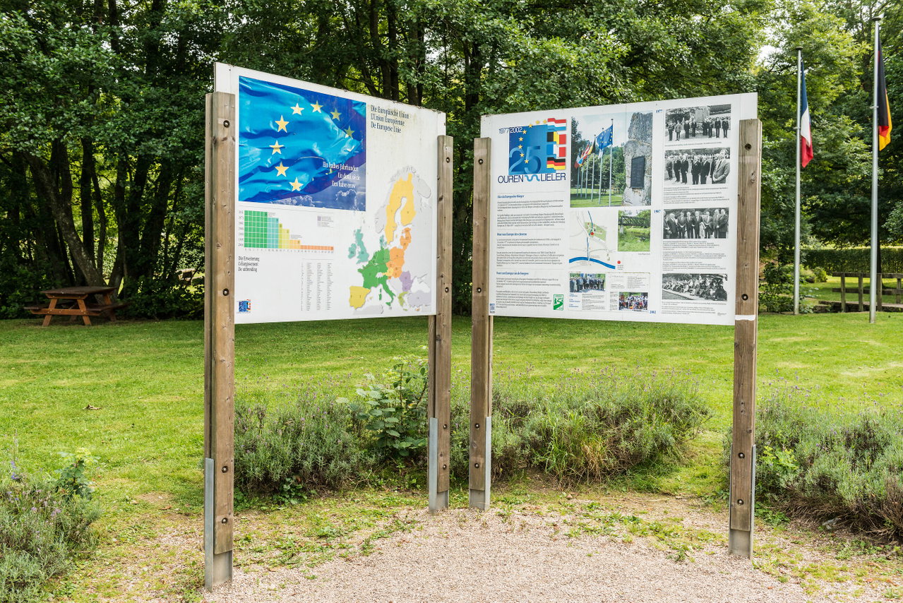 Gepflegte Parkanlage mit großem Gedenkstein, Infotafeln und mehreren Fahnen zwischen hohen, dicht belaubten Bäumen. Ein heller Kiesweg führt durch die grüne Anlage und vermittelt eine ruhige, einladende Atmosphäre.