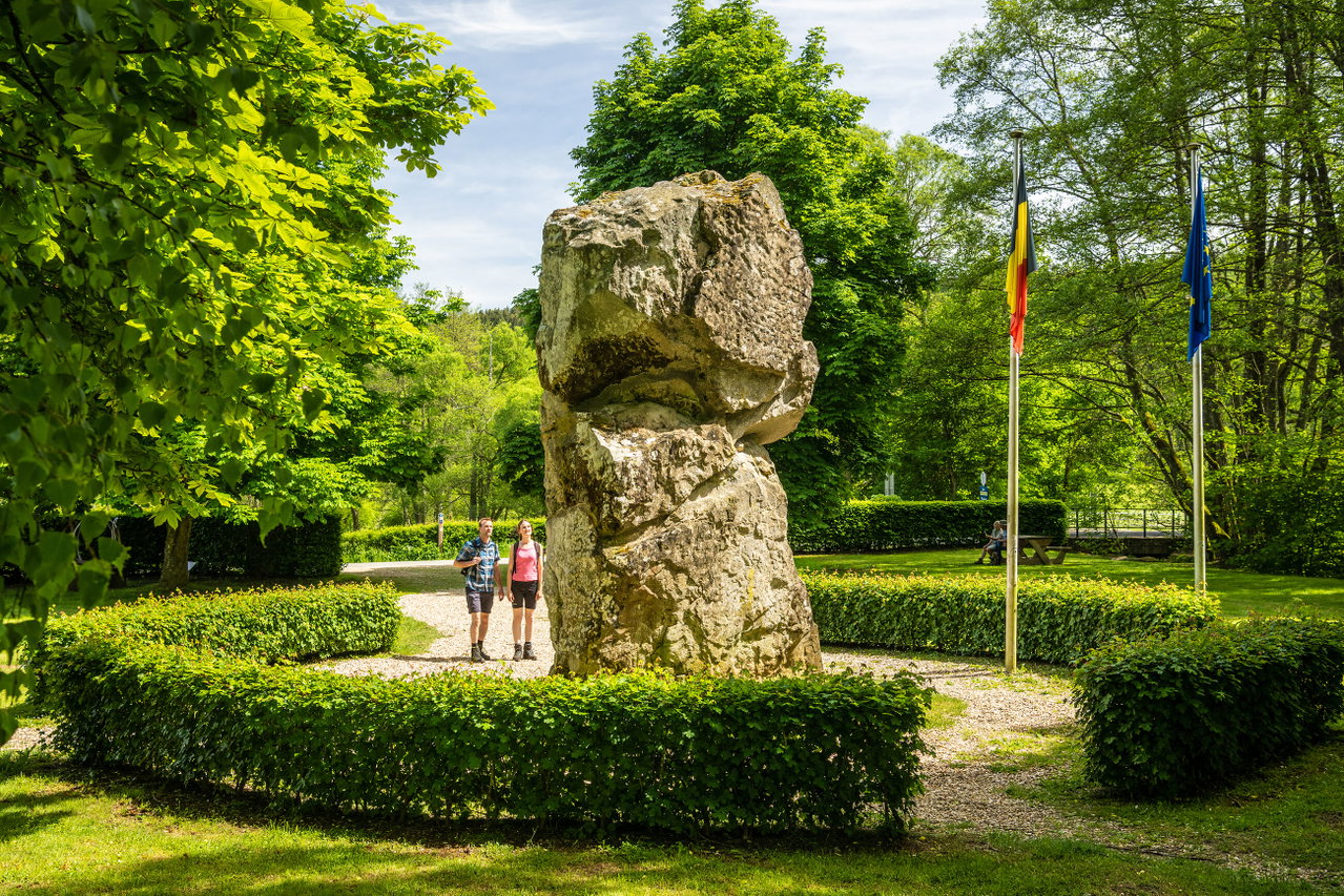 Großer Findling in einer gepflegten Parkanlage mit geschnittenen Hecken, umgeben von dichtem Grün. Rechts stehen die Flaggen von Deutschland und der Europäischen Union, während zwei Personen auf einem Kiesweg vorbeispazieren.