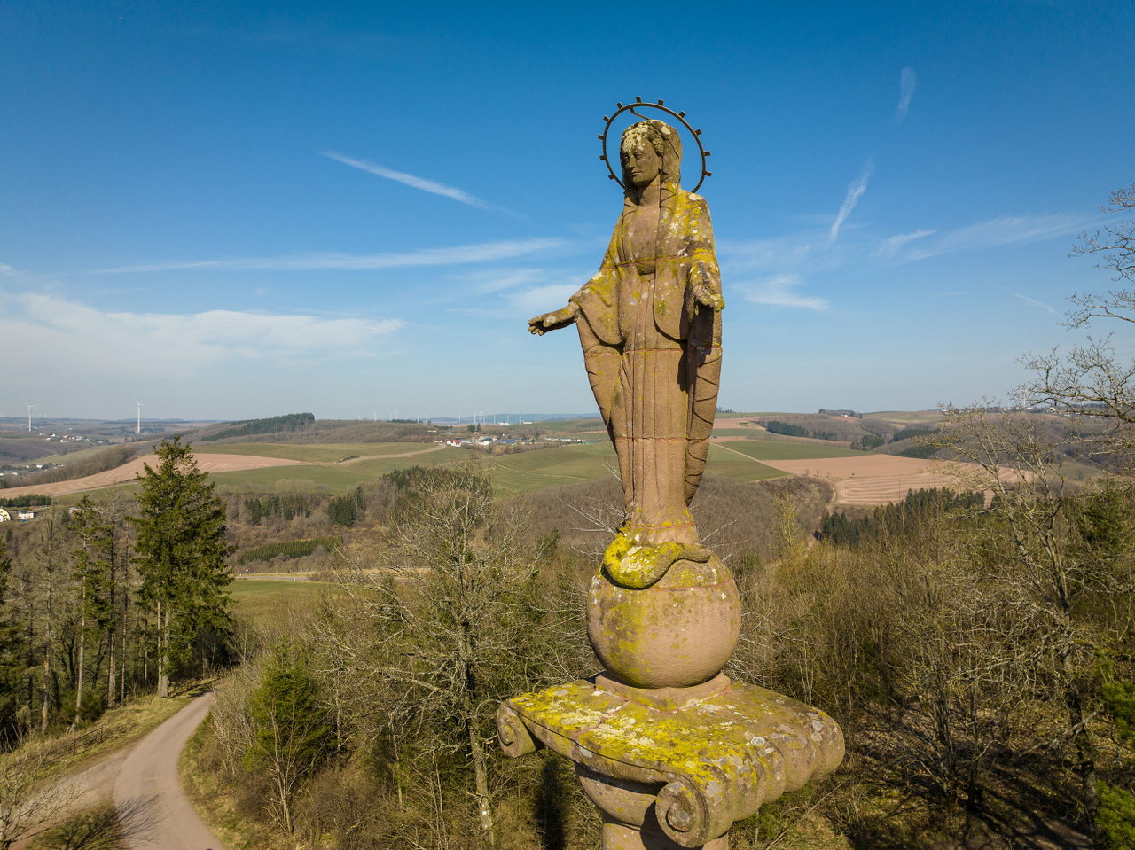 Die Mariensäule steht auf einem Hügel vor weitem Blick über das grüne Tal mit kleinen Ortschaften, Wiesen und bewaldeten Hängen unter blauem Himmel.