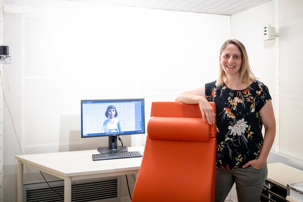 Silvia Kober steht im Psychologie-Labor an einen roten Sessel gelehnt, links im Bild ein Monitor, auf dem ein weiblicher Avatar zu sehen ist