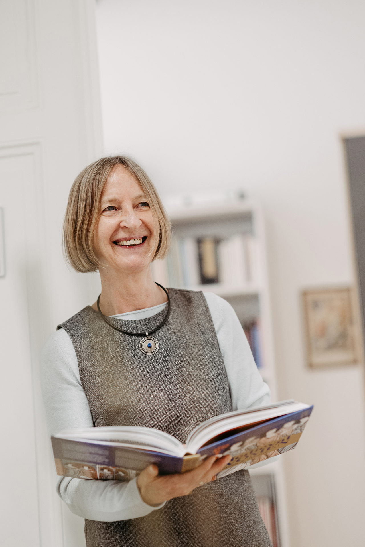 Brigitte Spreitzer-Fleck lehnt mit einem Buch in der Hand an der Wand und lächelt