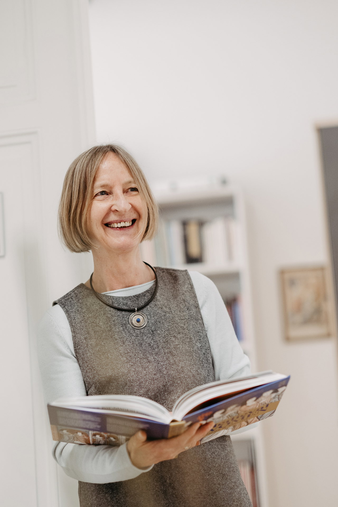 Brigitte Spreitzer-Fleck lehnt mit einem Buch in der Hand an der Wand und lächelt