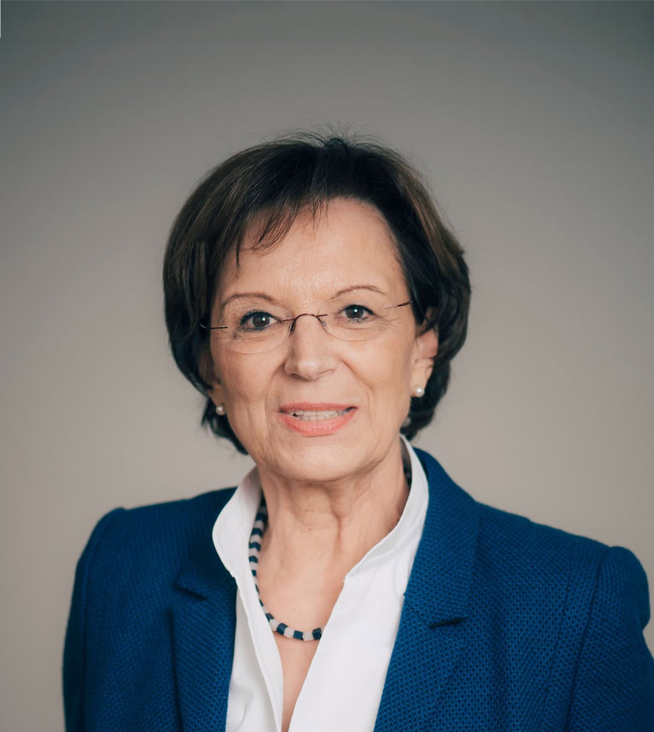 Porträt von Emilia Müller in blauem Blazer und weißer Bluse. Die Frau mit dem brünetten Kurzhaarschnitt trägt außerdem eine blau-weiße Perlenkette und eine runde Brille.