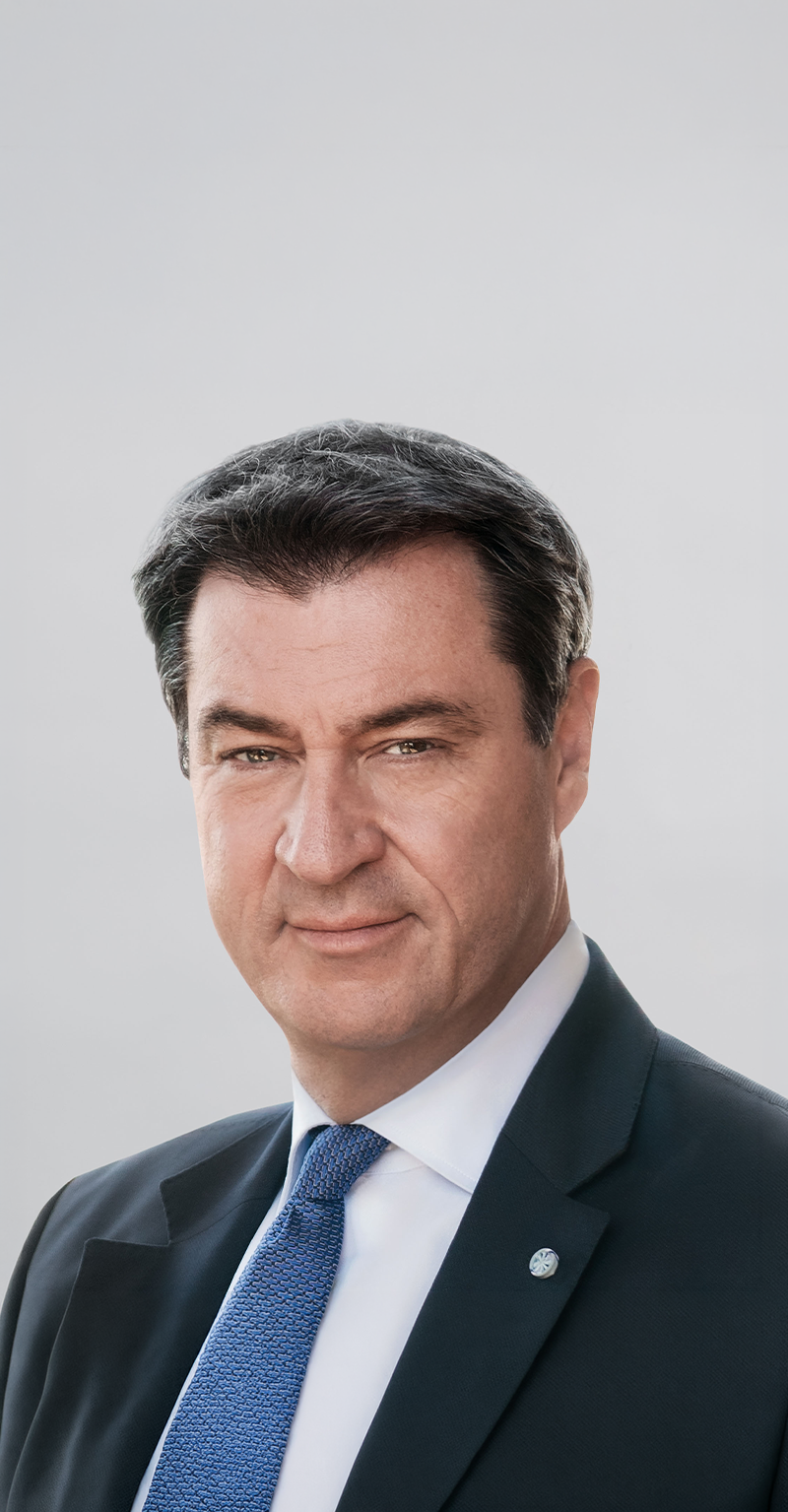 Porträt von Doktor Markus Söder vor hellem Hintergrund. Der Ministerpräsident trägt einen Anzug mit blauer Krawatte, seine Haare sind kurz und schwarz.
