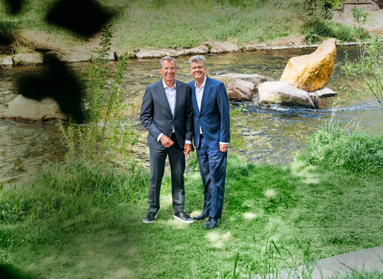 Die Brüder Christoph und Georg Wellendorff stehen in Anzügen nebeneinander am Ufer eines Flusses vor Wiese und Steinen.