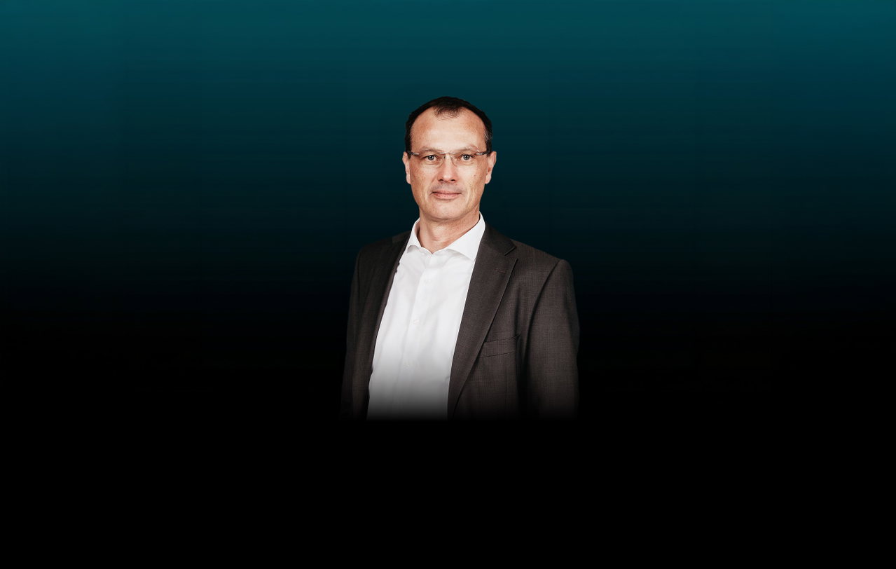 Freisteller von Peter Leitenmayer vor dunklem Hintergrund. Der Mann trägt einen braunen Anzug mit einem weißen Hemd sowie eine eckige Brille.