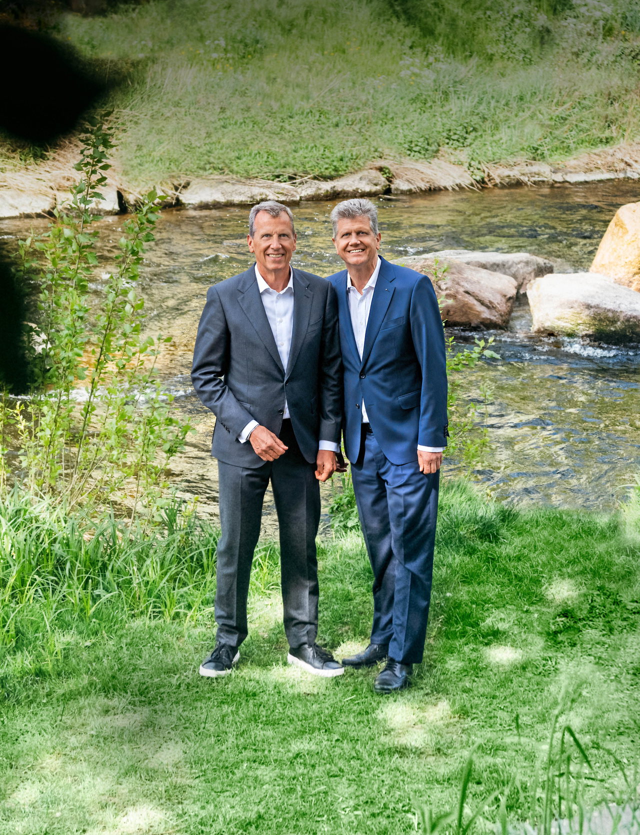 Christoph und Georg Wellendorff stehen im Anzug lächelnd nebeneinander am Ufer des Flusses Enz im Grünen.