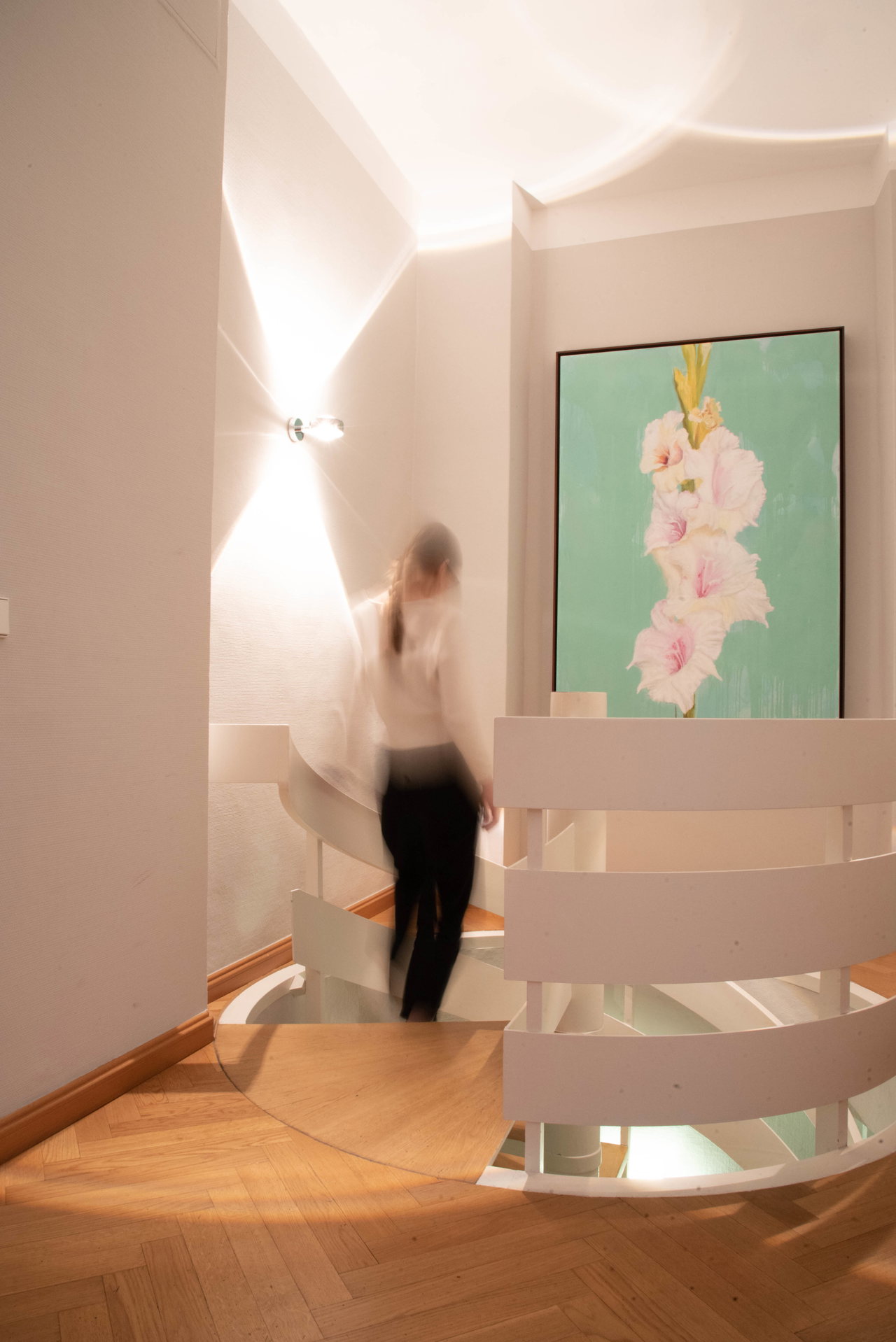 Person geht eine geschwungene weiße Treppe hinauf, an der Wand ein großes Gemälde mit rosa Blüten
