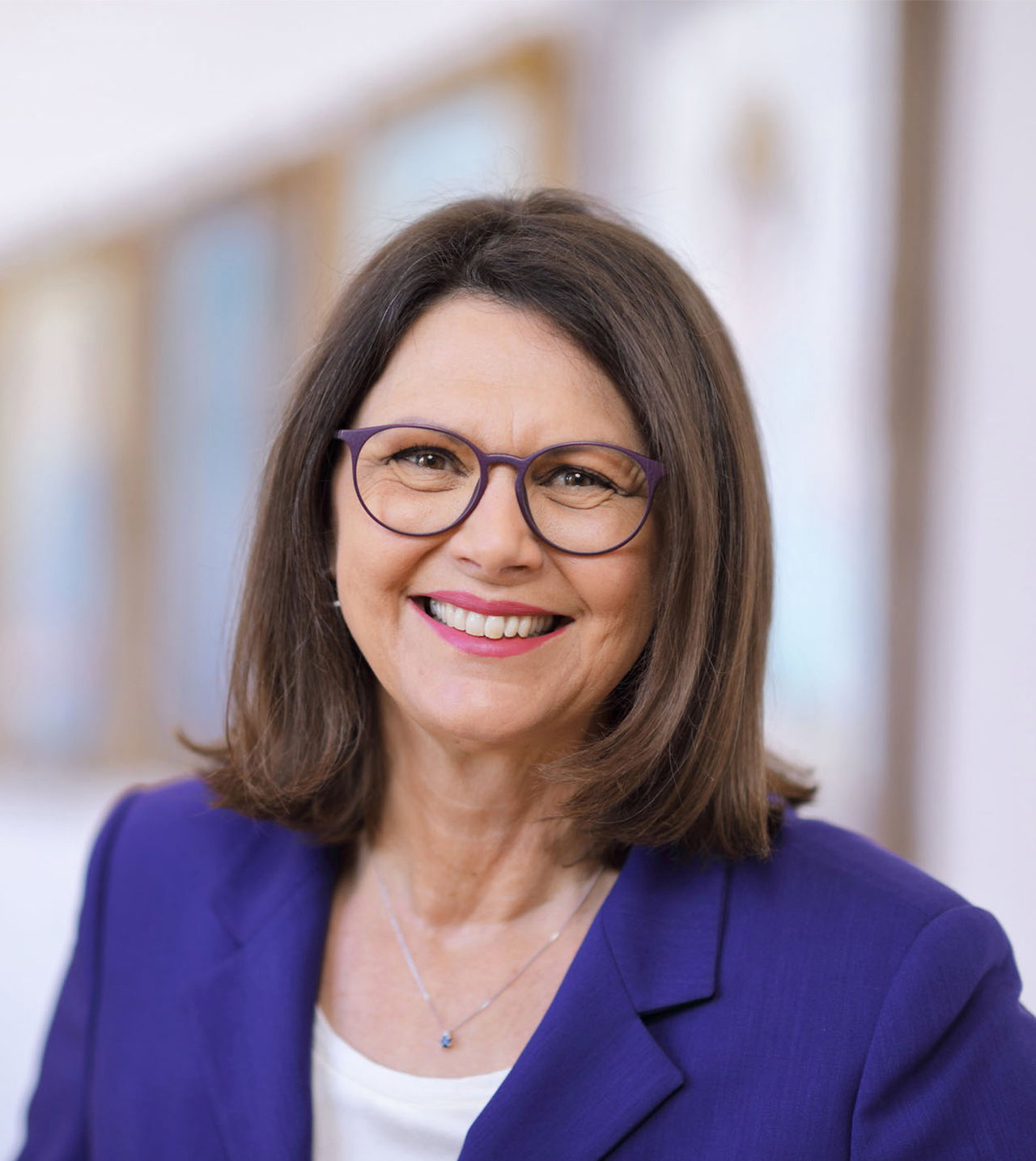 Porträt von Ilse Aigner in dunkelblauem Blazer, vor hellem Hintergrund. Die Frau trägt eine runde Brille, um ihren Hals eine zierliche Silberkette mit Anhänger. Sie hat schulterlange, braune Haare.