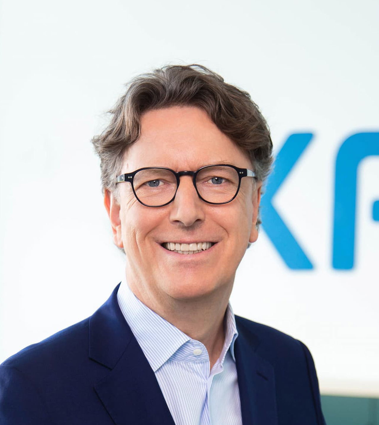 Porträt von Stefan Wintels in Anzug und mit runder Brille. Der Mann steht vor dem Logo der Kreditanstalt für Wiederaufbau: die blauen Buchstaben K, F und W auf weißem Hintergrund. 