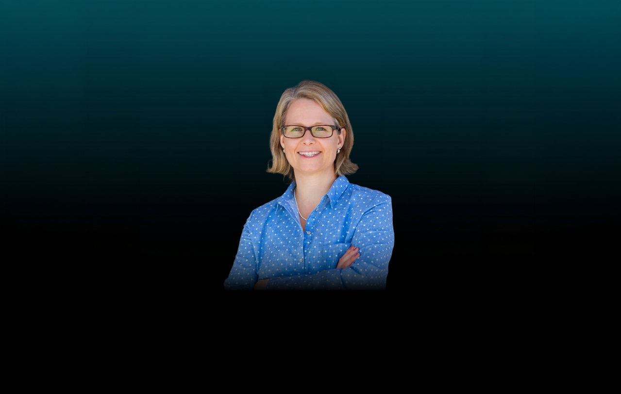 Freisteller von Christina Auer mit verschränkten Armen vor dunklem Hintergrund. Die Frau mit den schulterlangen Haaren trägt eine blaue Bluse mit weißen Punkten und eine dunkle, eckige Brille.