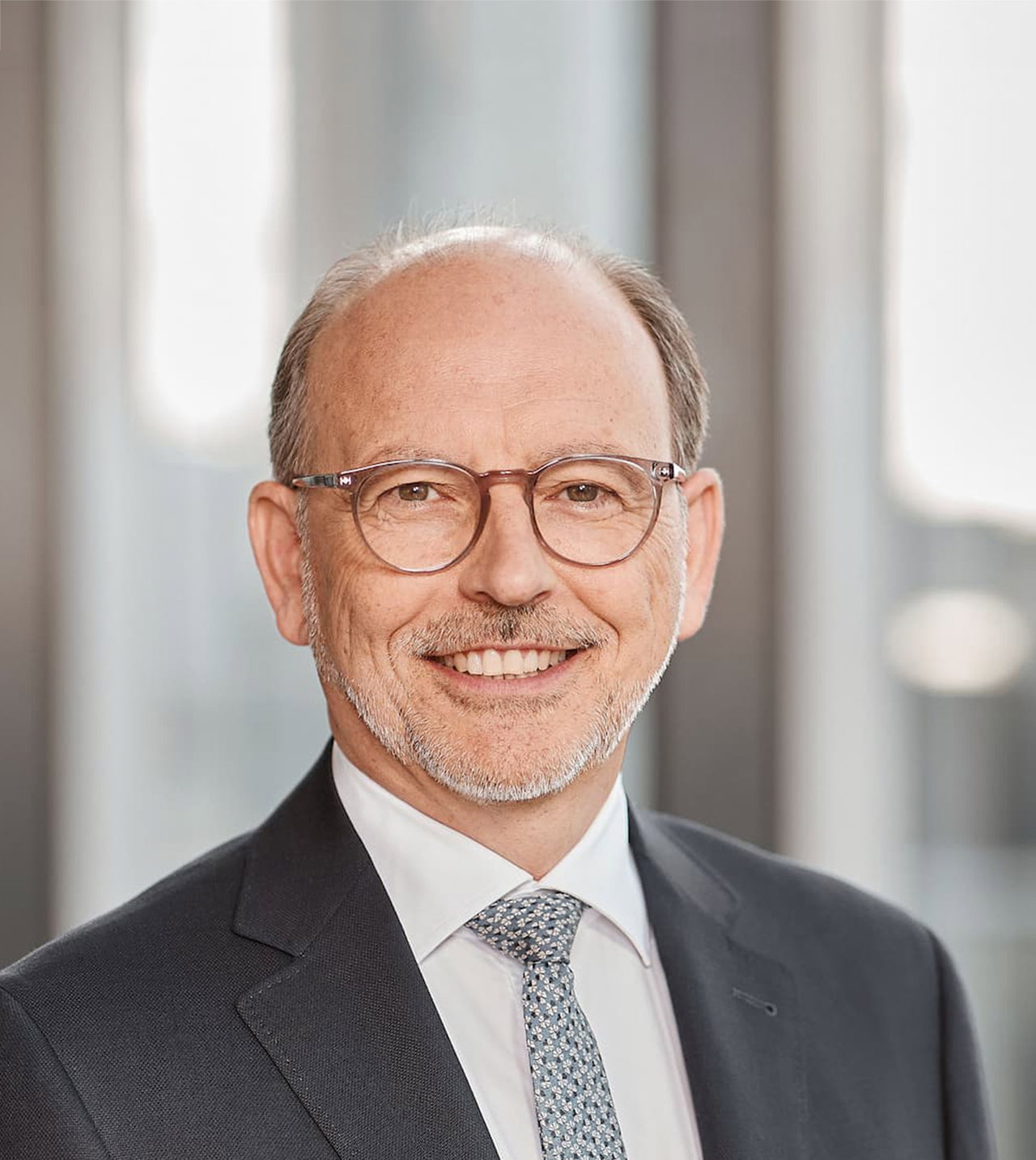 Porträt von Thomas Groß vor hellem Hintergrund. Er trägt einen dunkelgrauen Anzug, eine gemusterte Krawatte und eine runde Brille. 