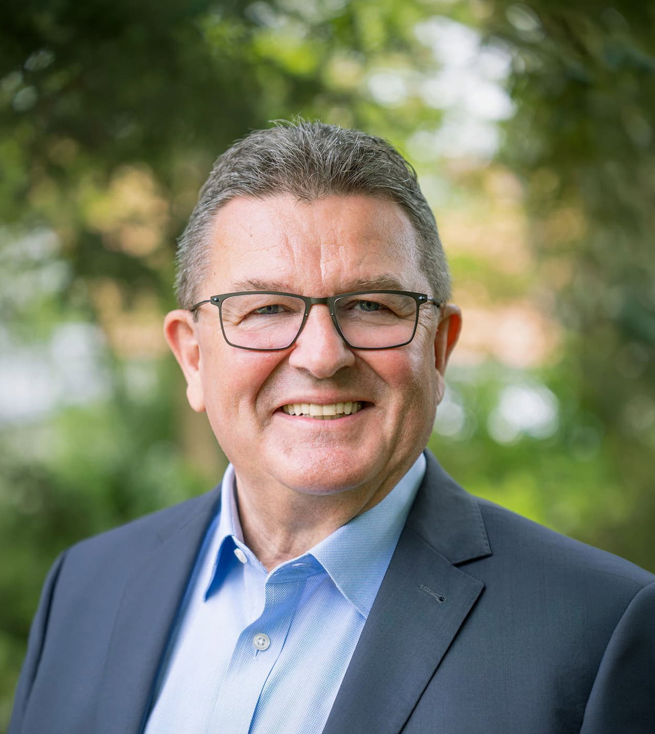 Porträt von Franz Josef Pschierer, im Hintergrund unscharfe grüne Blätter. Der grauhaarige Mann trägt einen Anzug und eine eckige, dunkle Brille.