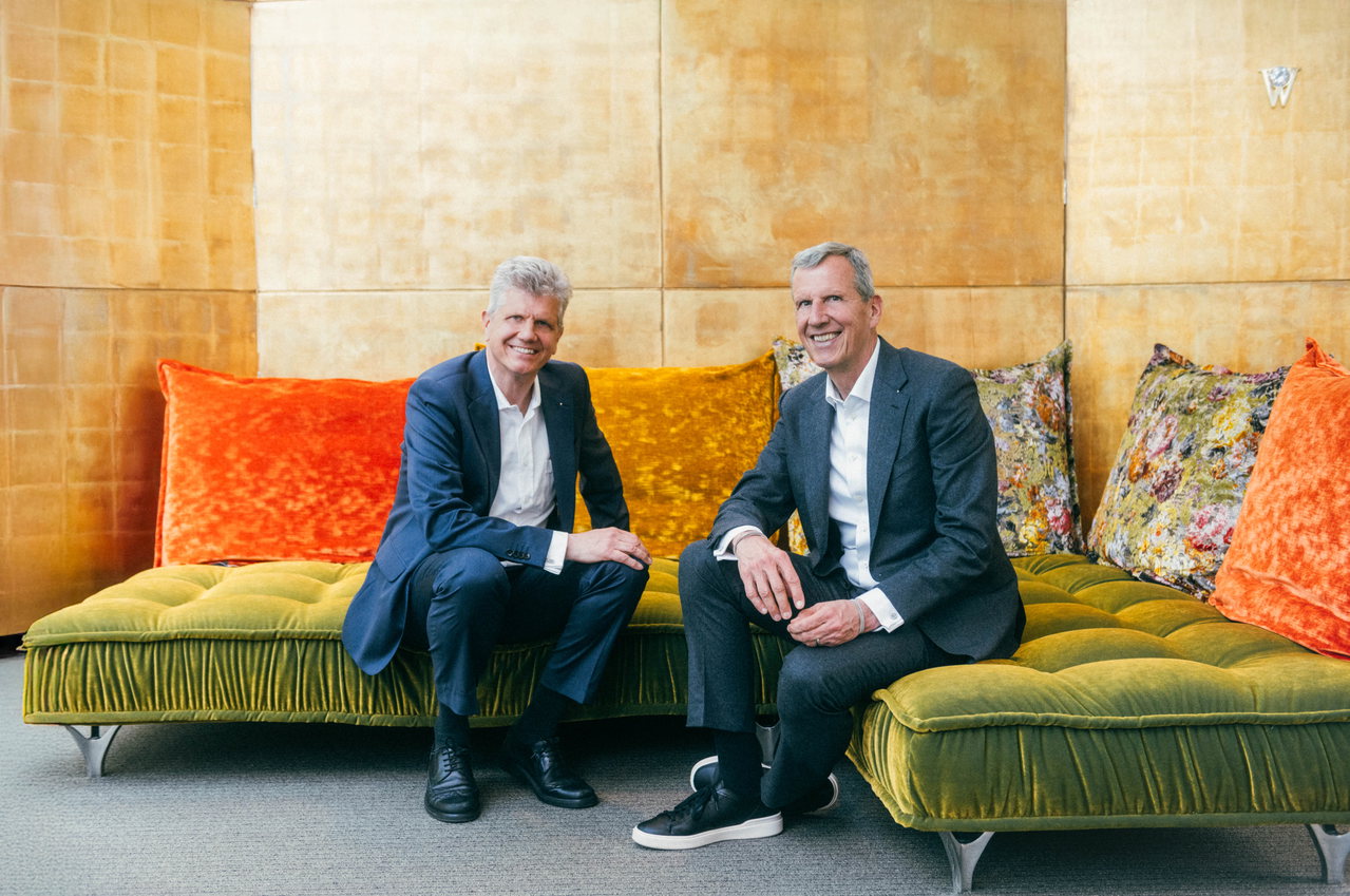 Georg und Christoph Wellendorff sitzen in Anzügen lächelnd auf einer grünen Polsterbank vor einer goldenen Wand.