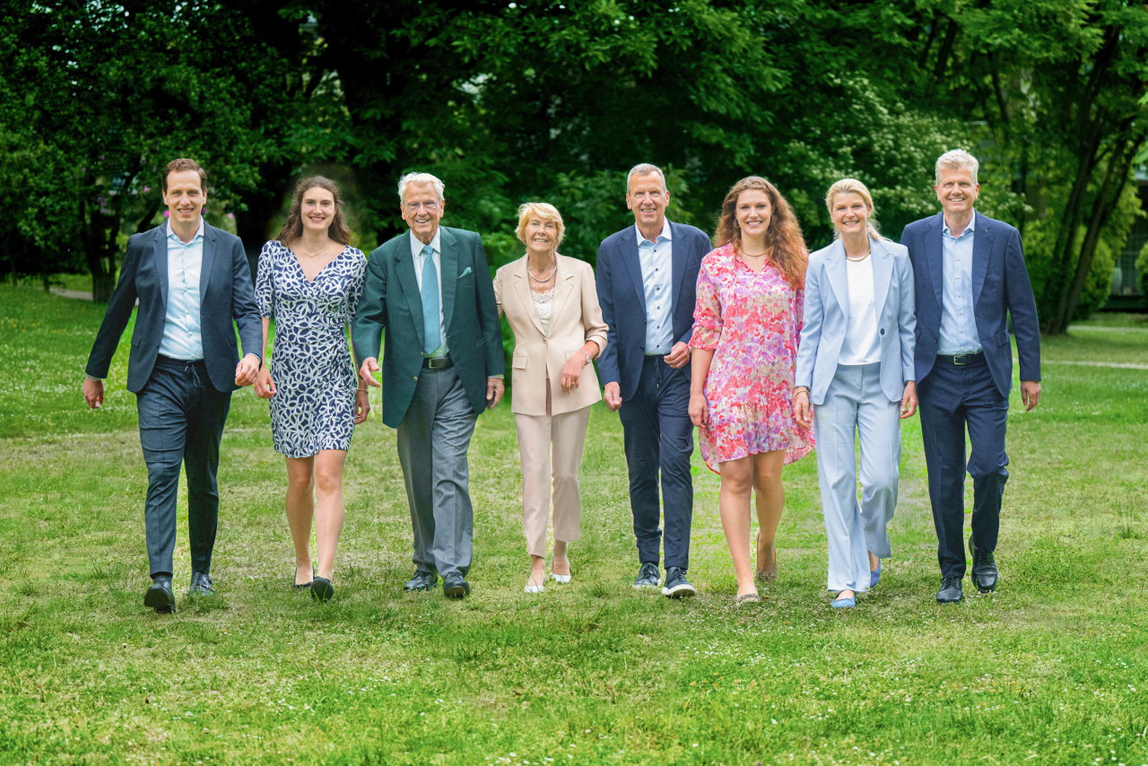 Acht Mitglieder der Familie Wellendorff, formell und leger gekleidet, gehen nebeneinander auf einer Rasenfläche vor Bäumen auf die Kamera zu.