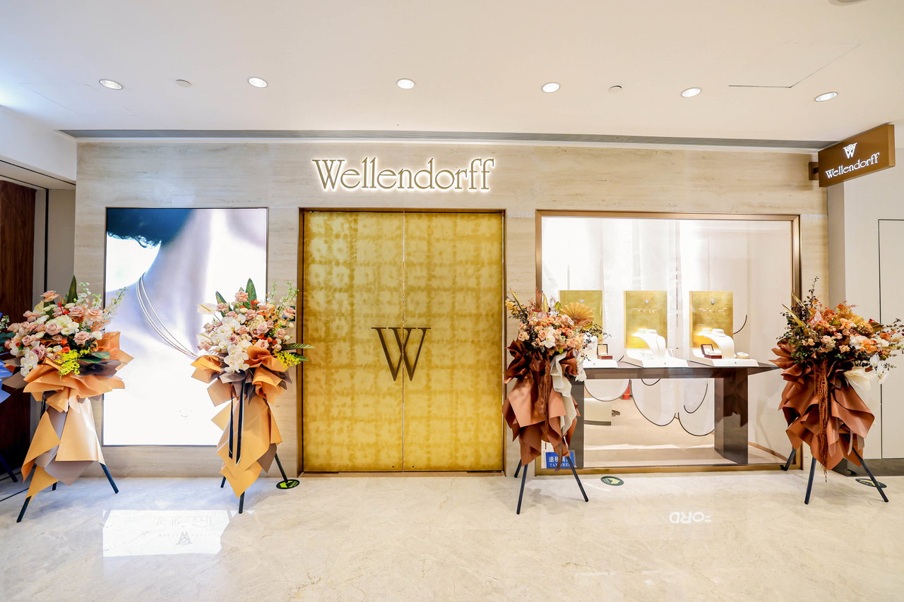 Innenansicht des Eingangs der Wellendorff-Boutique in der China World Mall in Peking mit goldener Tür, großem Markenlogo, Schaufensterdisplays und vier Blumengestecken auf Ständern.