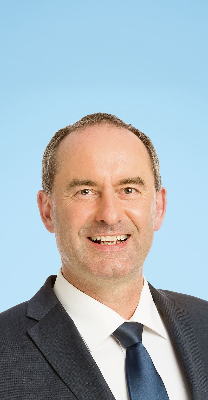 Porträt von Hubert Aiwanger vor hellblauem Hintergrund. Der kurzhaarige Mann trägt einen Anzug mit schwarzer Krawatte.