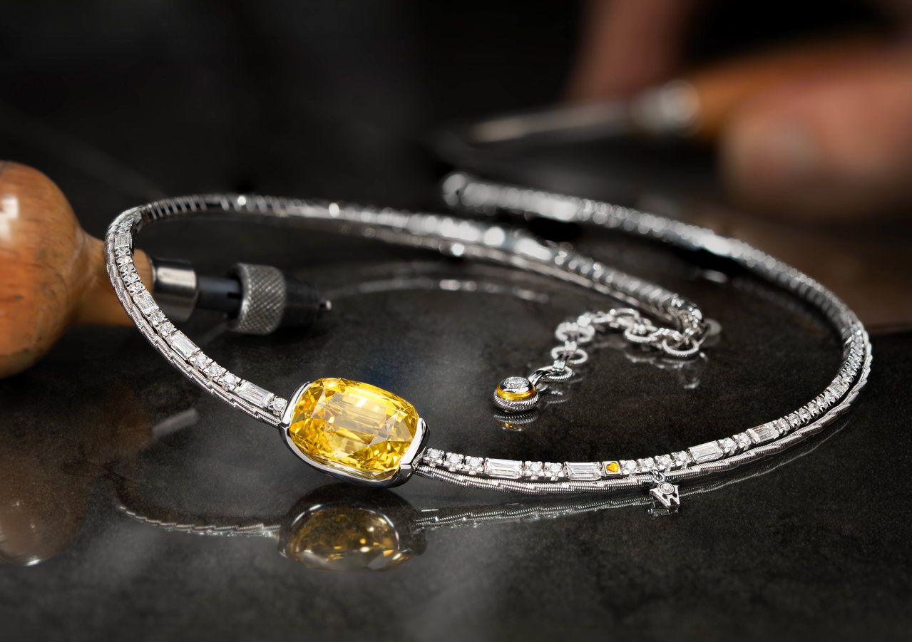 Wellendorff Collier SONNENSCHEIN. in Weißgold mit einem großen gelben Edelstein liegt auf einer dunklen Oberfläche vor Goldschmiedewerkzeugen.