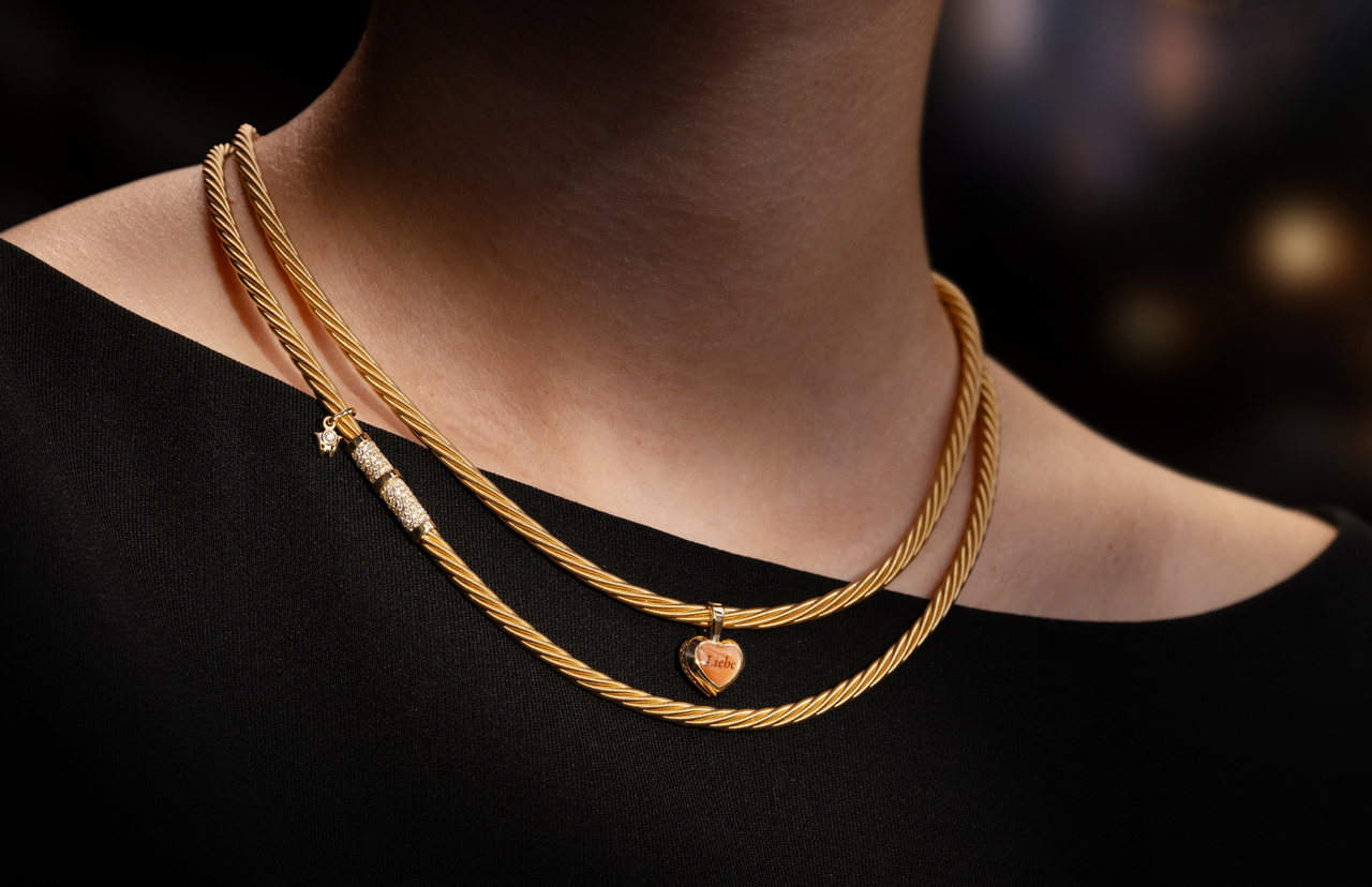 Eine Frau trägt ein zweireihiges goldenes Wellendorff-Collier mit graviertem Herzanhänger über einem schwarzen Oberteil.