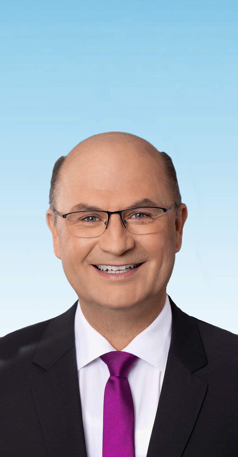 Porträt von Albert Füracker vor hellblauem Hintergrund. Der Mann ist im Anzug mit violetter Krawatte und eckiger Brille abgebildet. 