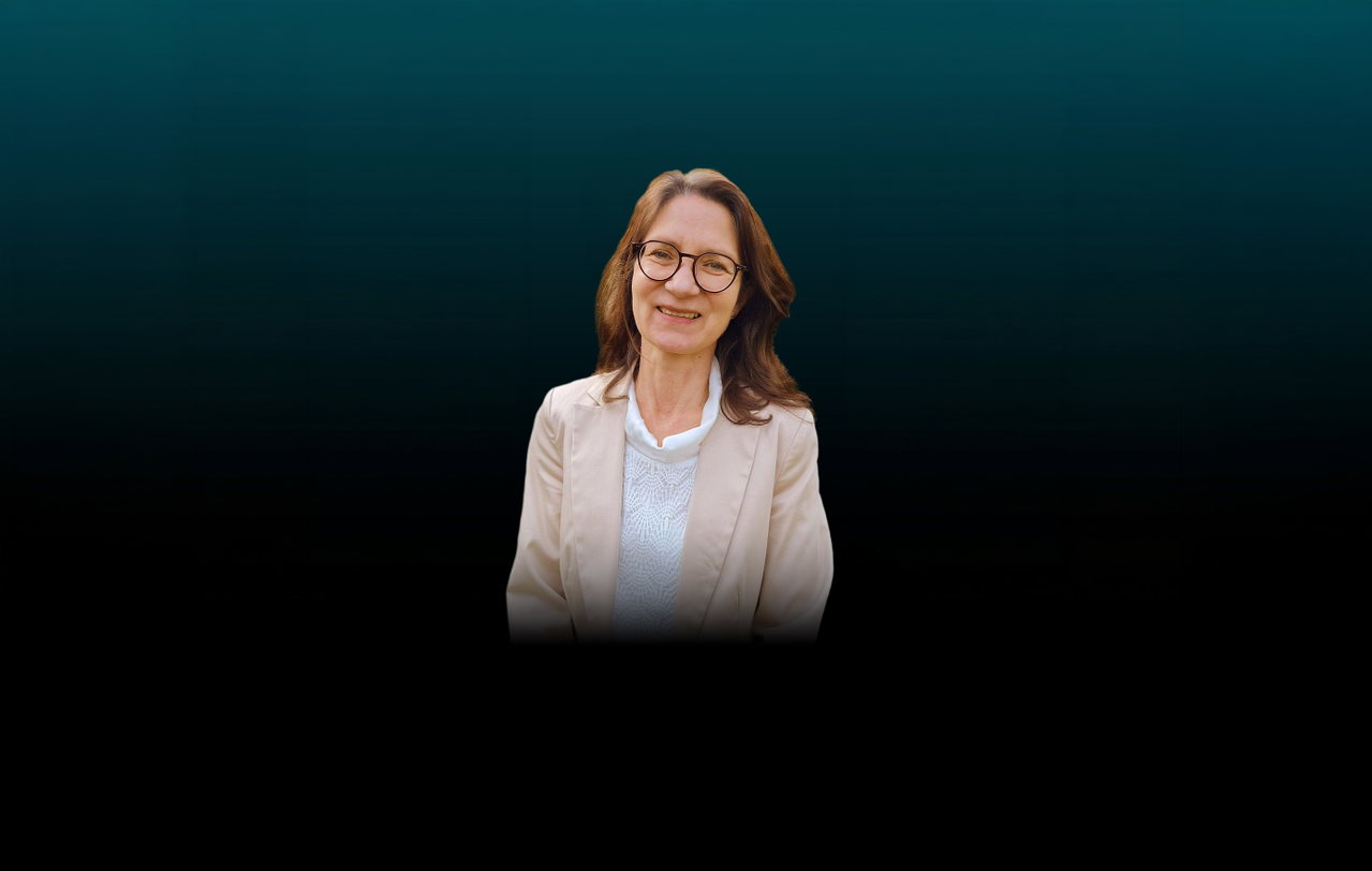 Freisteller von Sylvia Weingart vor dunklem Hintergrund. Die Frau hat schulterlanges, braunes Haar. Sie trägt eine weiße Bluse, dazu einen hellen Blazer und eine runde Brille.
