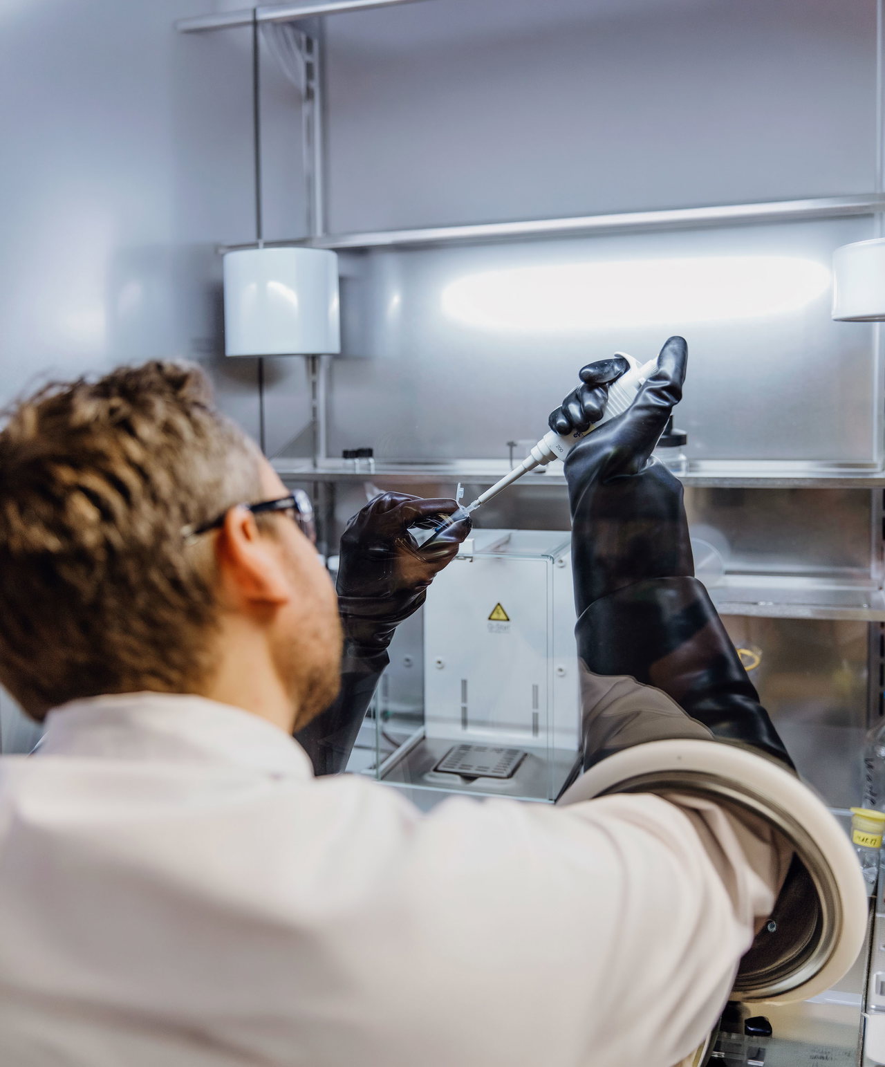 Abgebildet ist ein Mann im weißen Kittel von hinten. Er steht vor einer metallischen Laborwand und führt eine mechanische Pipette in eine Plastikhülse ein. Der Mann trägt schwarze Schutzhandschuhe sowie eine Schutzbrille.