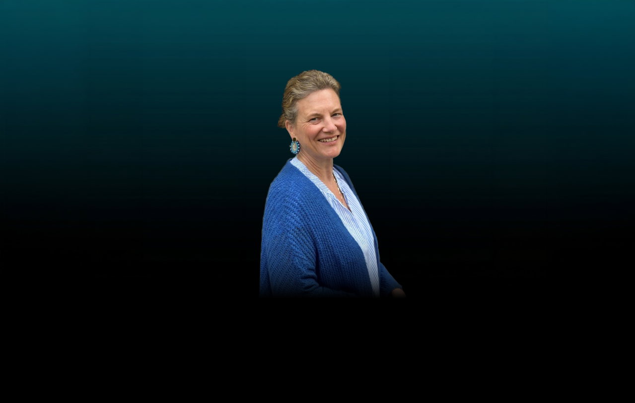 Freisteller von Annalena Köhler vor dunklem Hintergrund. Die Frau trägt eine dunkelblaue Wolljacke und eine hellblau gestreifte Bluse. Ihr Haar ist nach hinten gebunden.