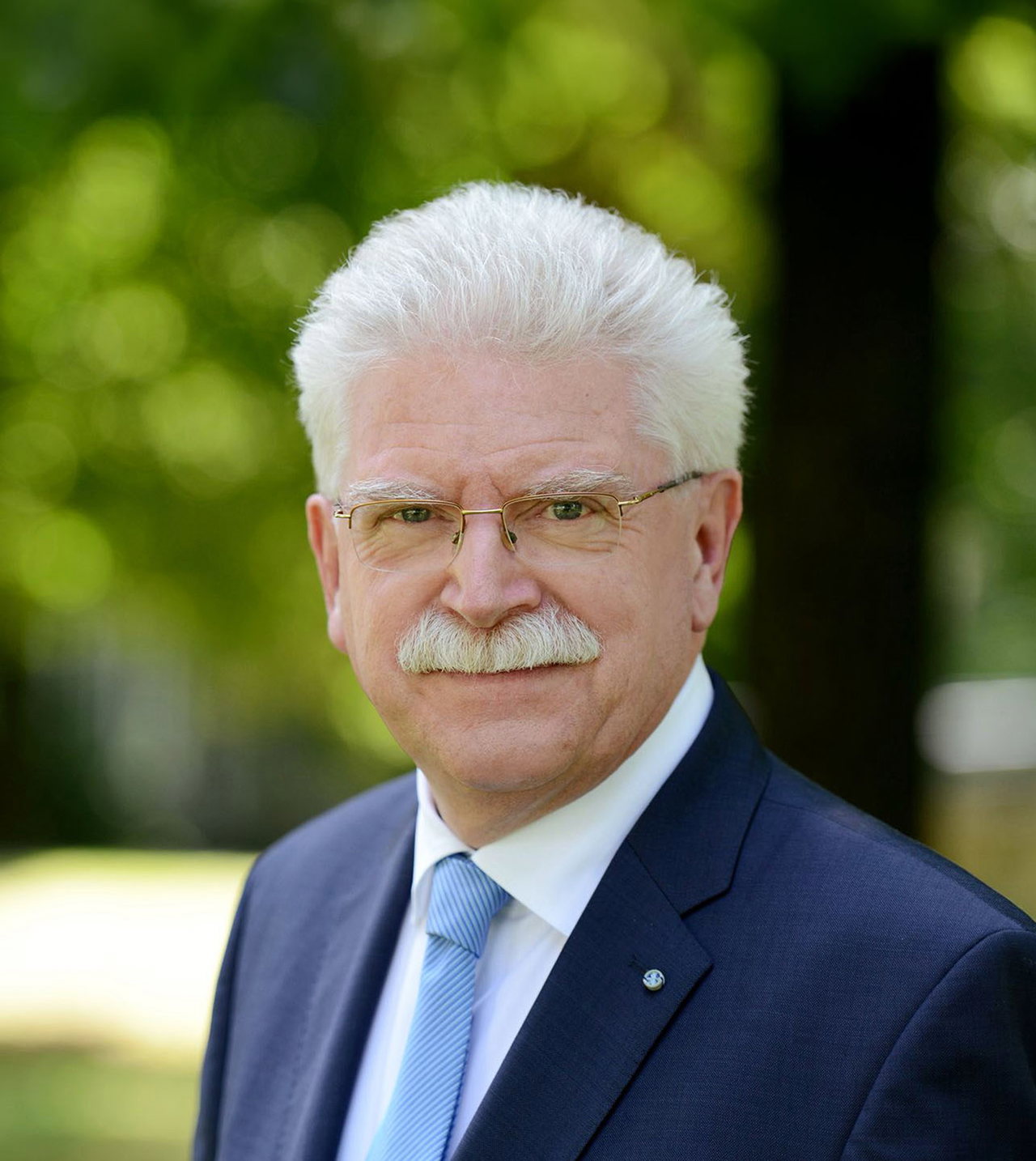 Porträt von Martin Zeil in dunkelblauem Anzug. Im unscharfen Hintergrund ist ein Baum zu erkennen. Der Mann hat einen weißen Schnauzer, weiße Haare und trägt eine Brille.