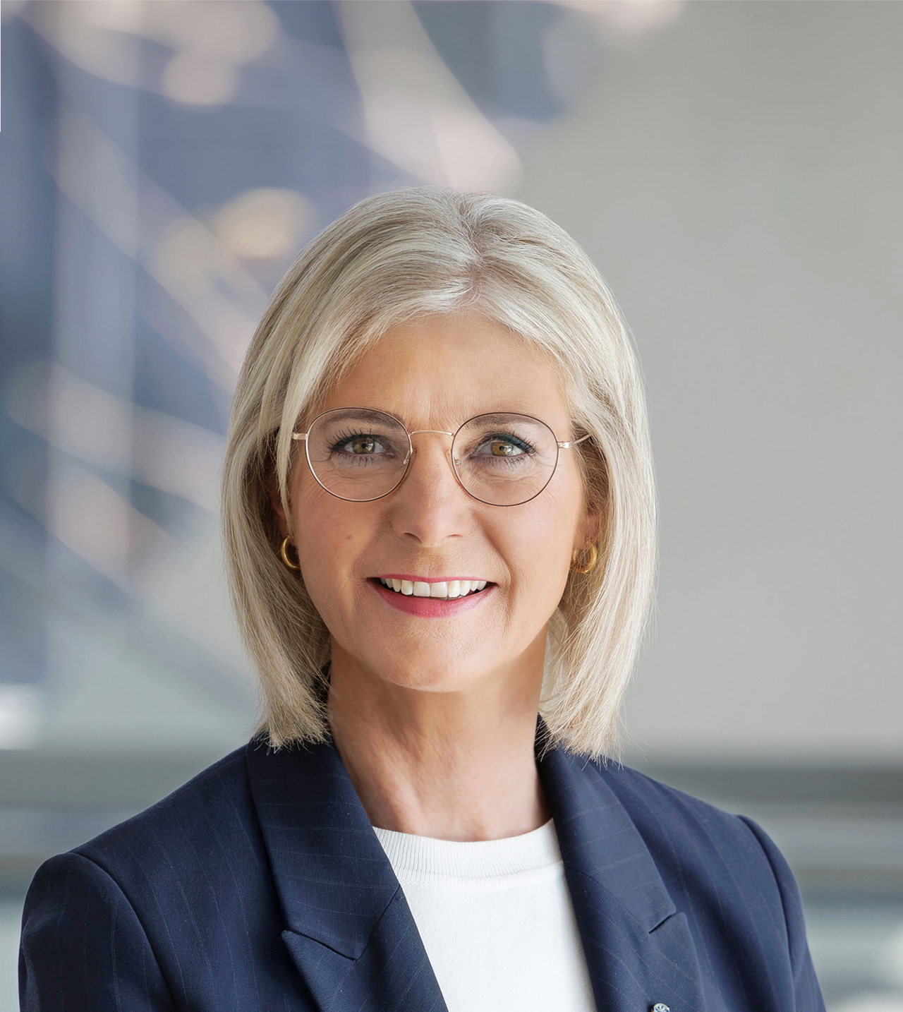 Porträt von Ulrike Scharf vor hellem Hintergrund. Die Frau trägt blonde Haare, eine runde Brille und zarte Kreolen. 