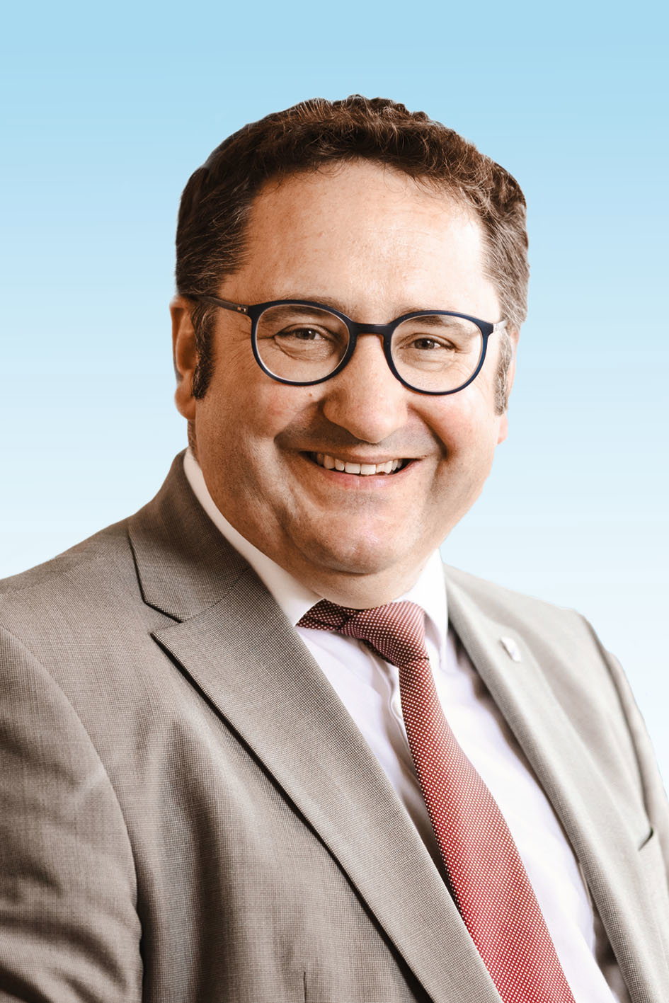 Porträt von Tobias Gotthardt vor hellblauem Hintergrund. Der Mann ist in beigem Anzug, rostbrauner Krawatte und runder Brille abgebildet. Er trägt kurzes braunes Haar. 