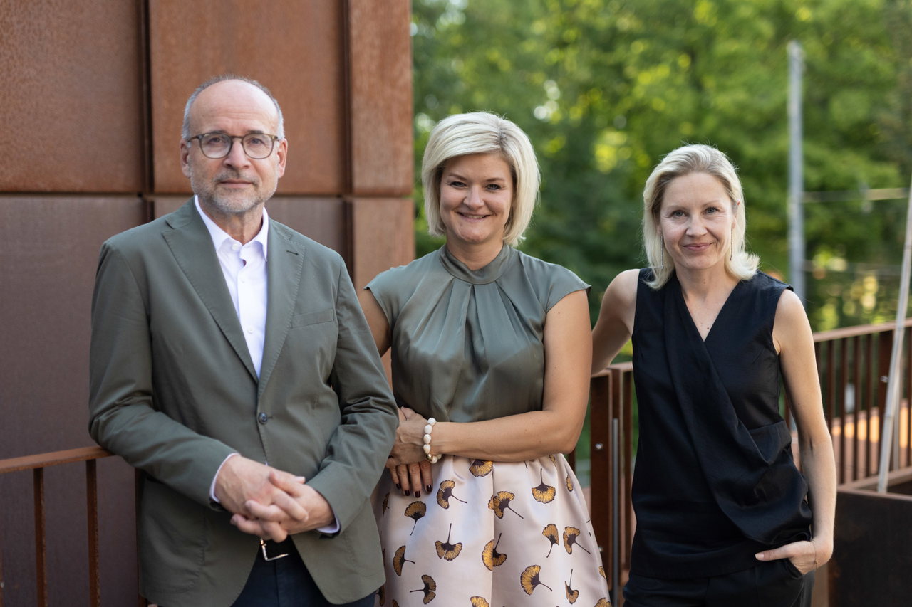 Agenturgründer Dr. Markus Schönmann und die storyboard-Geschäftsführerinnen Marie Bressem und Christine Fehenberger stehen nebeneinander im Freien vor einer Wand aus Cortenstahl und grünen Bäumen