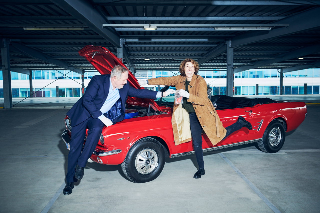 Mann im Anzug und Frau im Mantel posieren in einem Parkhaus an einem roten Oldtimer-Cabrio mit geöffneter Motorhaube