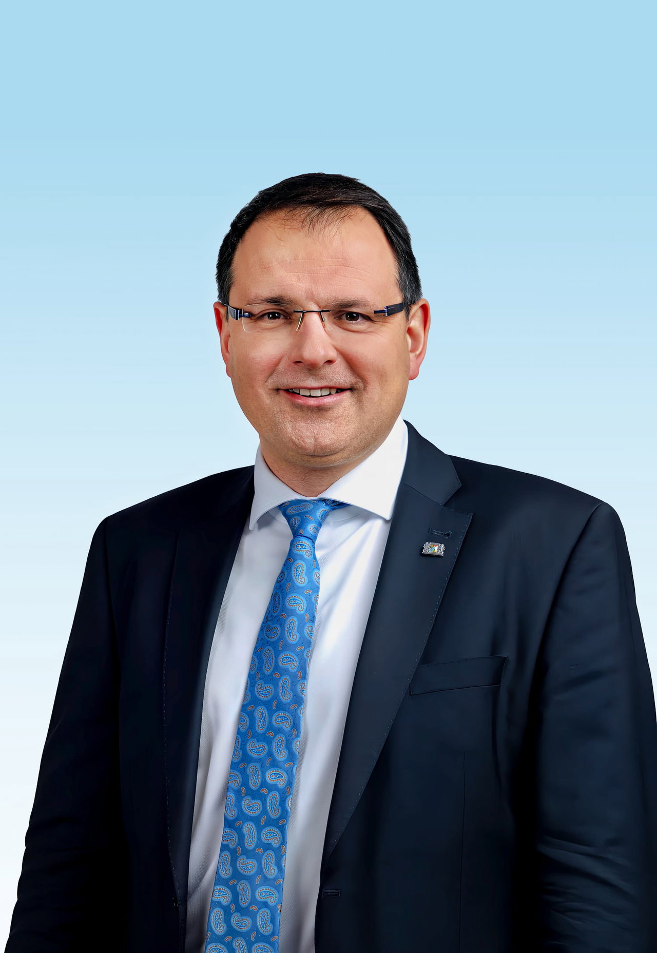 Porträt von Martin Schöffel im Anzug. Er trägt eine blaue, gemusterte Krawatte und eine eckige Brille. Der Hintergrund ist hellblau.