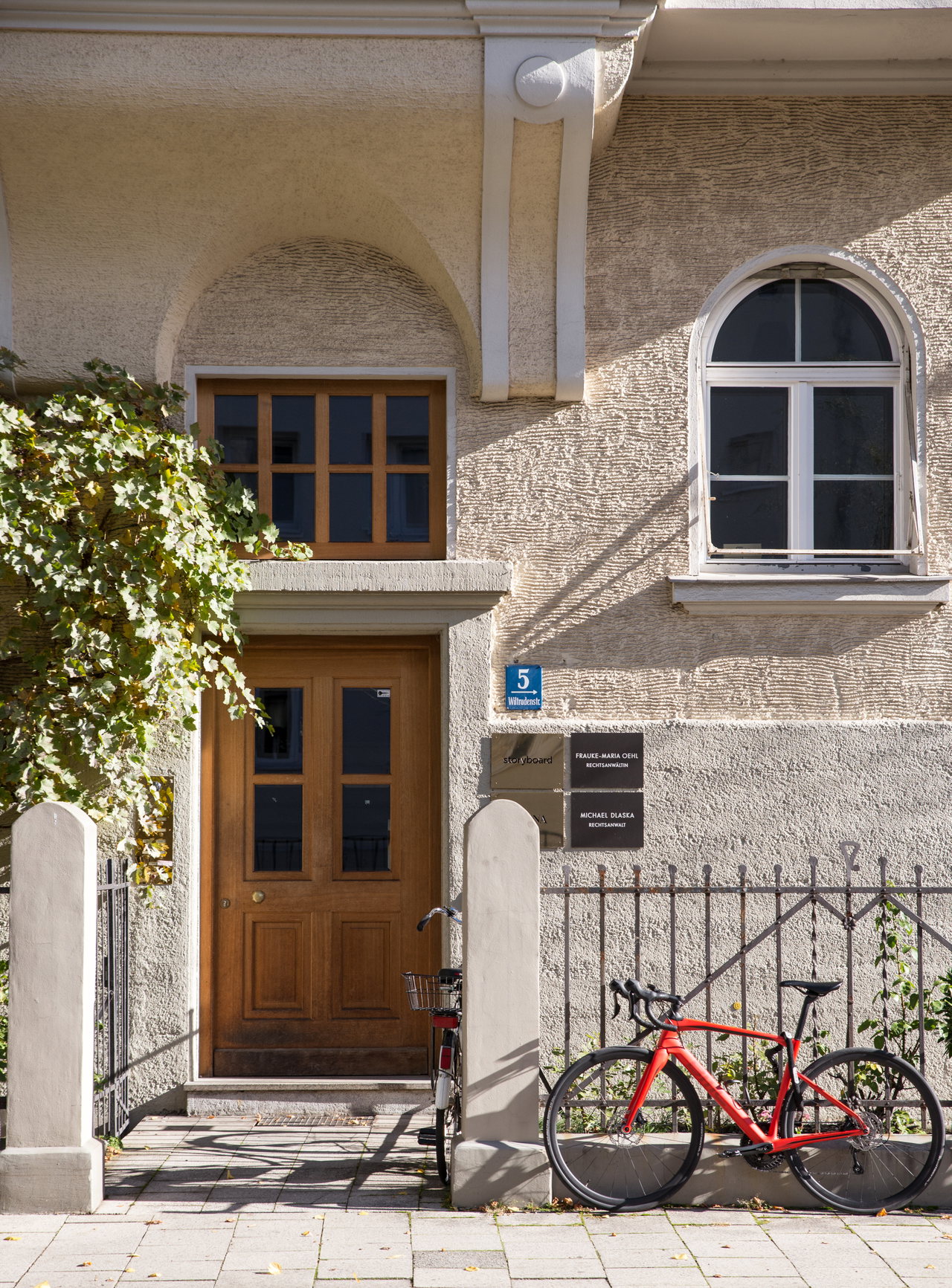 Eingang des Altbaus in der Wiltrudenstraße: Holztür mit Rundbogen, davor ein rotes Rennrad am gusseisernen Zaun