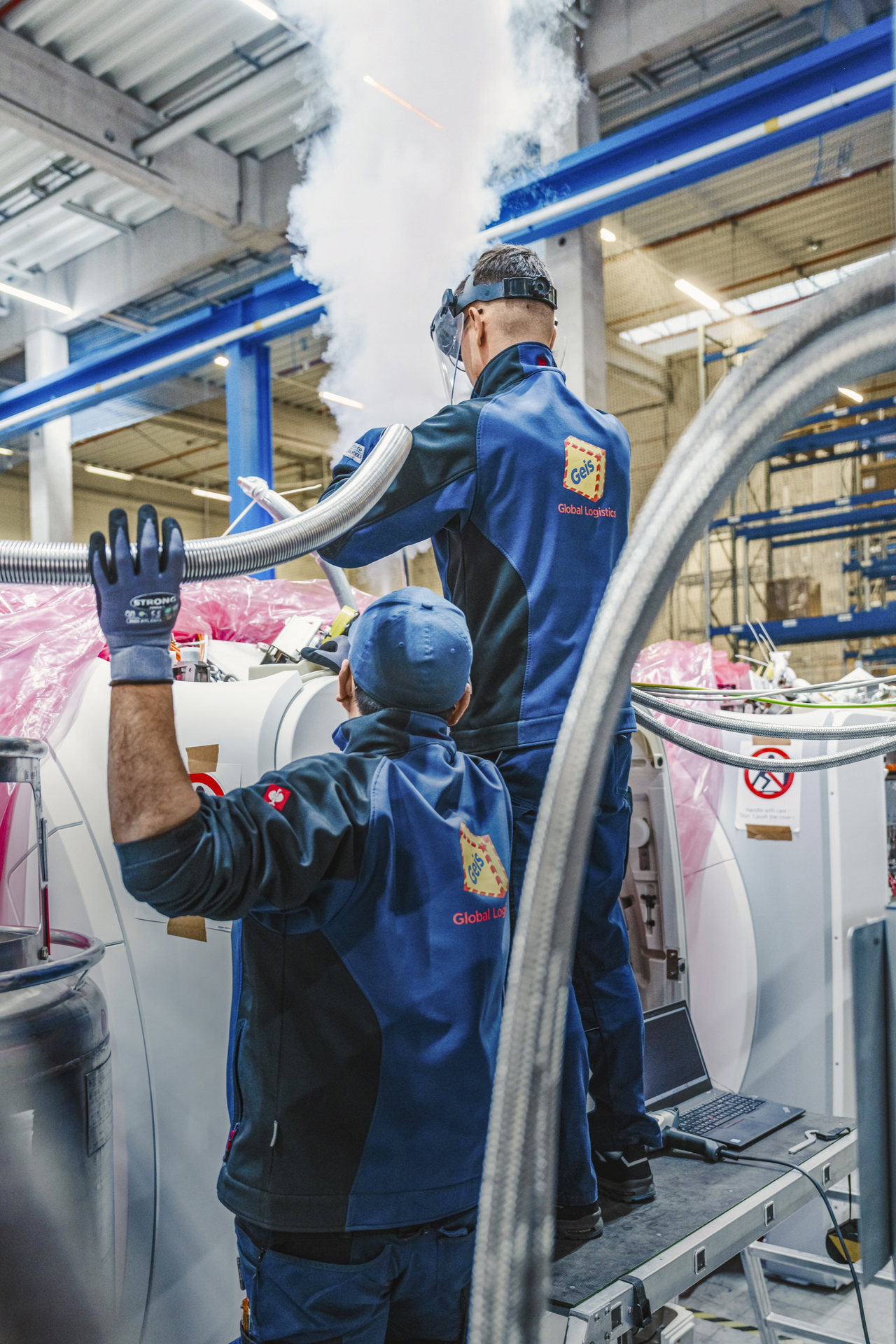 Zwei Techniker befüllen ein MRT mit flüssigem Helium, dabei entsteht eine Dampfwolke